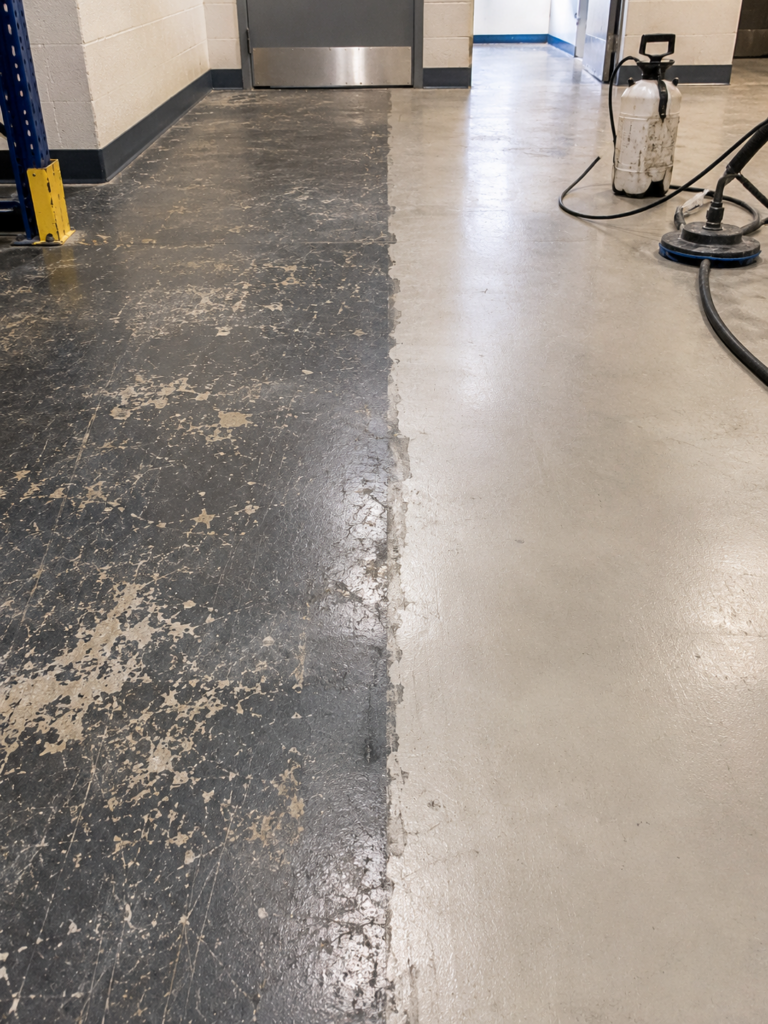Warehouse hallway with a split floor: left half in chipped black coating, right half polished gray concrete; cleaning equipment in the corner.