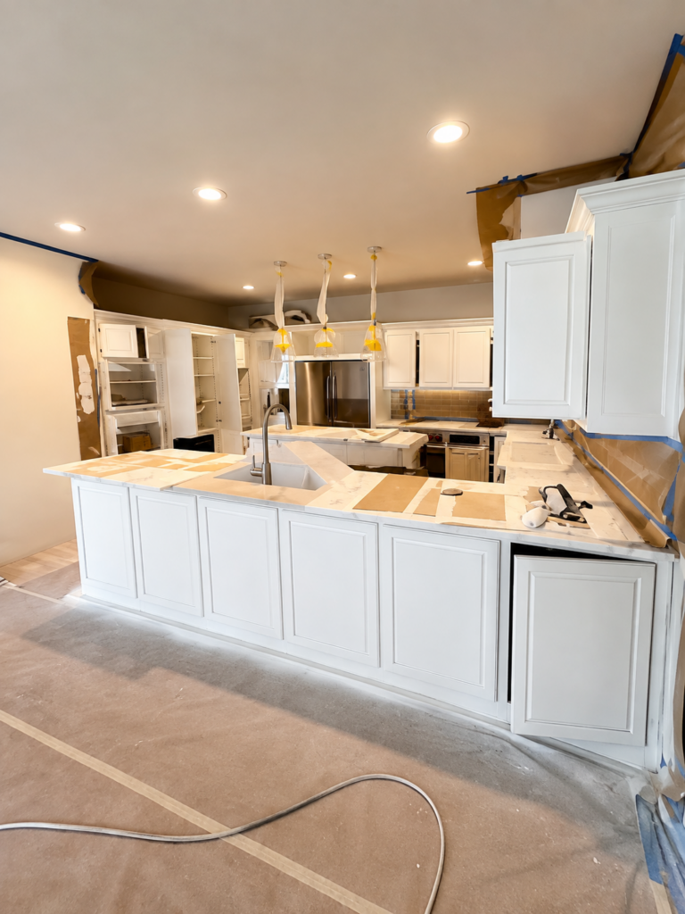 White kitchen undergoing renovation with an island, stainless steel appliances, and taped-off edges awaiting finish. 본 Pending fixtures and protective coverings visible.