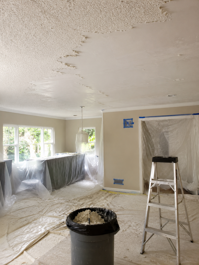 Room under renovation with plastic coverings over countertops and windows, a ladder, and a bucket of plaster on the floor.