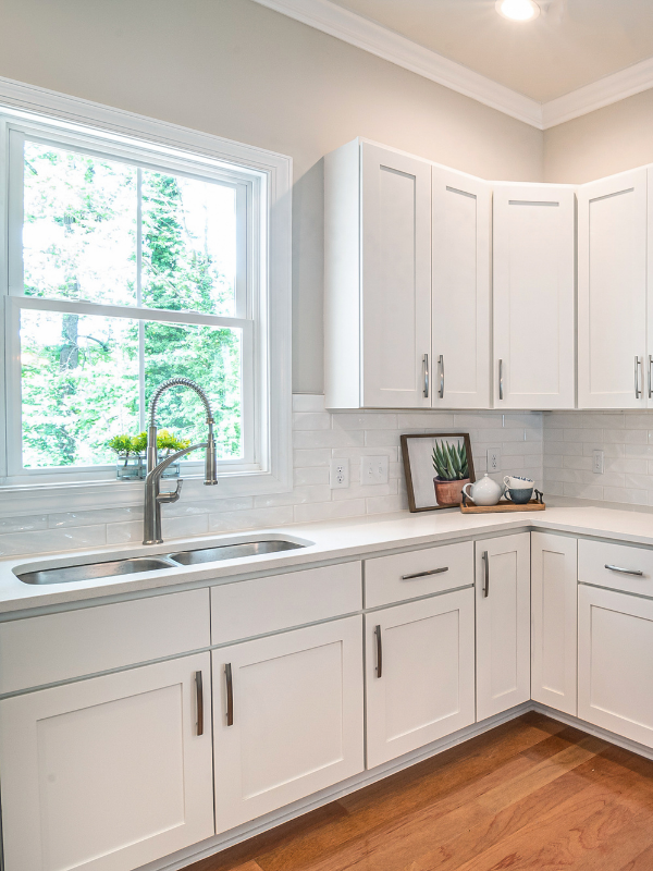 Bright white kitchen with a stainless steel sink under a window, white cabinets, and light wood flooring.