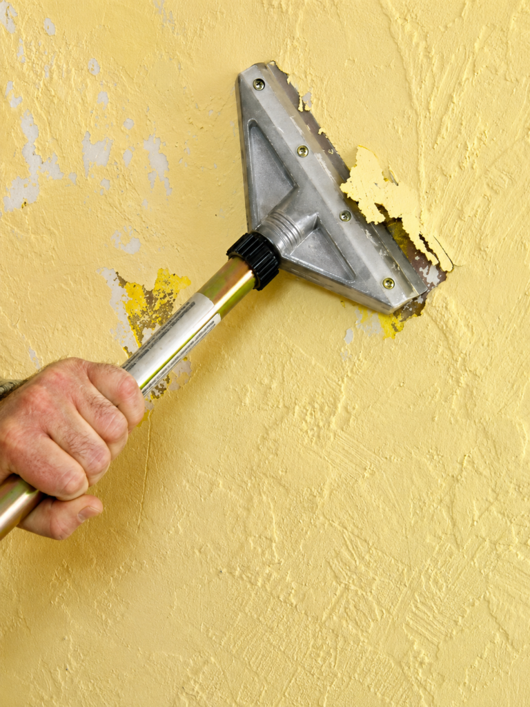 Person uses a triangular metal scraper to remove yellow paint from a textured wall, hand gripping a long handle.